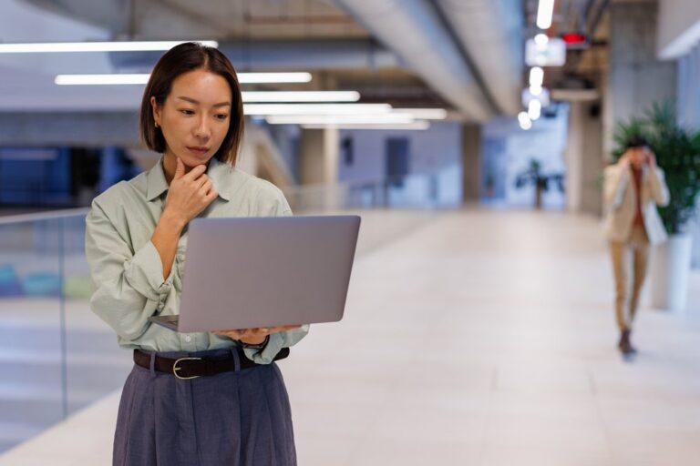 Businesswoman pausing thoughtfully over a laptop in an office walkway, representing comparing Philippines staffing partners